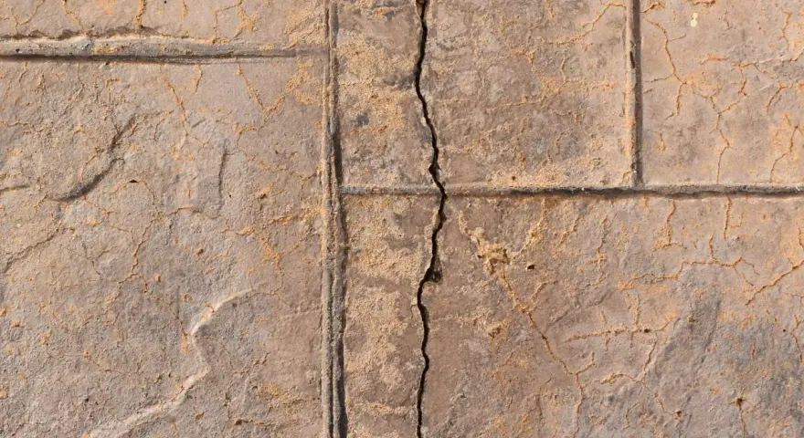 A prominent crack runs through a stamped concrete slab foundation, revealing sand and smaller fissures.