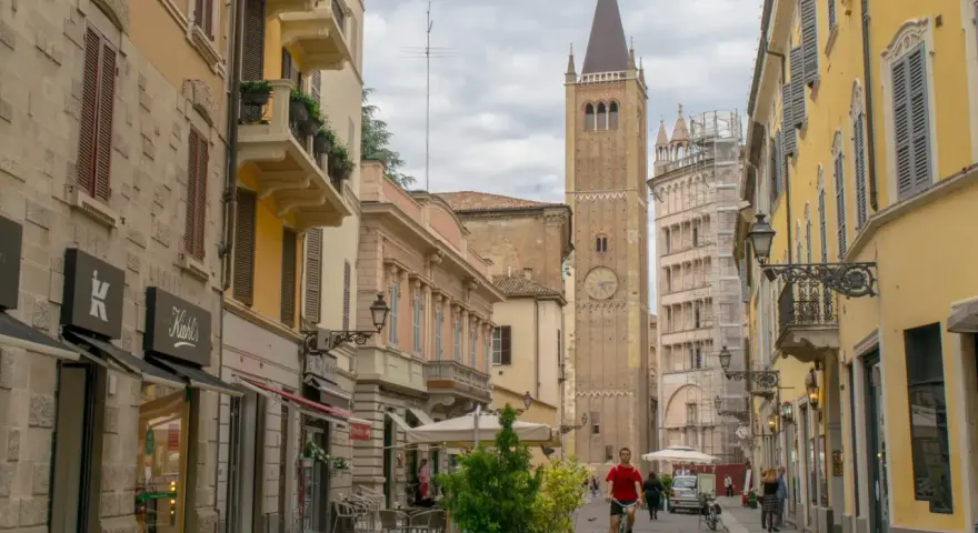 A man cycles down a street in Parma, Italy, with a tall clock tower in the background. This view captures the essence of a journey from Bologna to Parma.