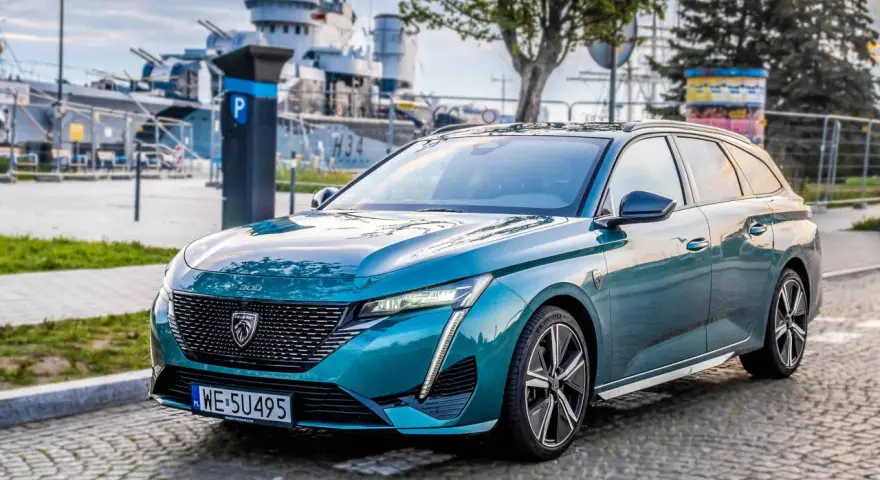 Turkusowe kombi Peugeot 308 zaparkowane na bruku, w tle okręt wojenny.