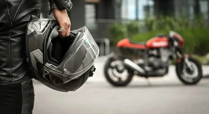Motociclista in pelle tiene un casco, pronto per misurare la circonferenza della testa per un acquisto perfetto. Moto rossa sfocata sullo sfondo.