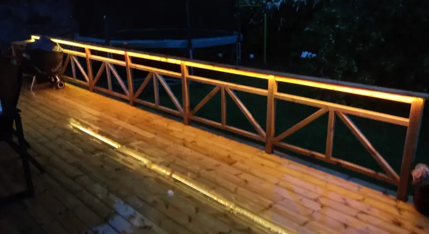 Träaltan med staket upplyst av varma decklights. En grill står på altanen.