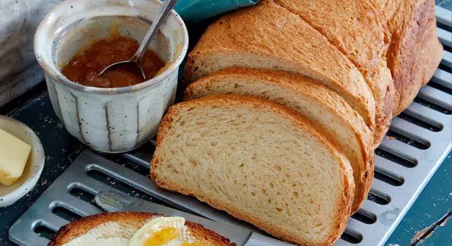 Ein Laib **sauerteig Toastbrot** ist aufgeschnitten. Daneben stehen Marmelade, Butter und ein Messer mit einem Stück Brot, das bestrichen ist.