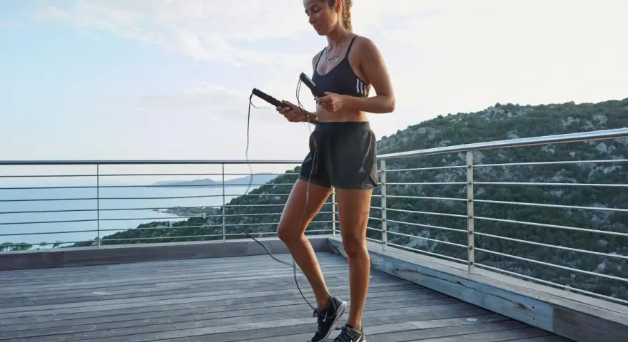 Femme en tenue de sport tenant une corde à sauter, prête pour un exercice en plein air avec vue sur la mer.