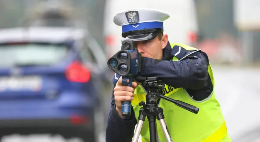 Policjant w kamizelce odblaskowej mierzy prędkość samochodu za pomocą radaru. Trwa dozór policyjny na drodze.