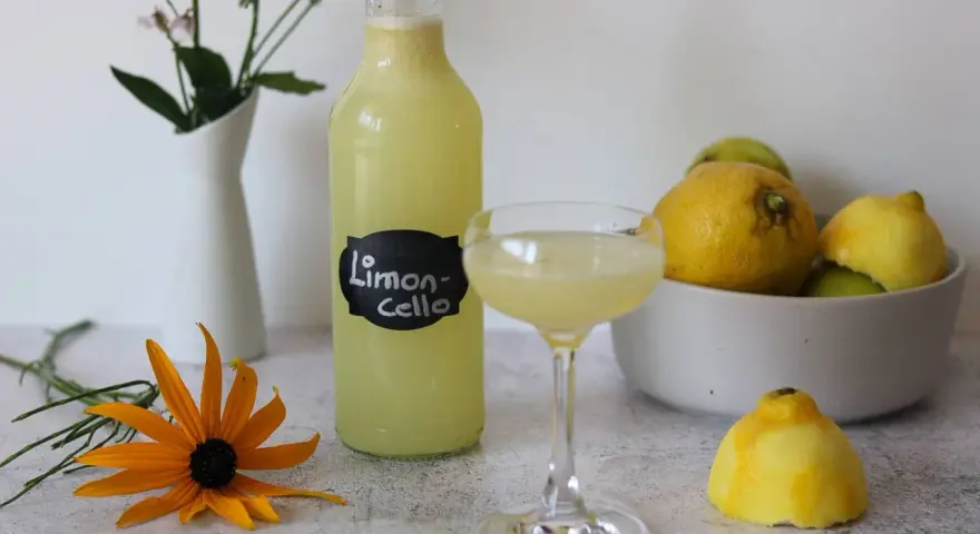 Erfrischendes Limoncello Eis, serviert in einem Glas, neben einer Flasche und frischen Zitronen. Ein gelber Sonnenhut blüht daneben.