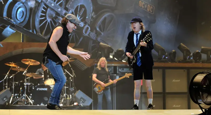 Brian Johnson singt, Angus Young spielt seine ACDC Gitarre. Im Hintergrund eine Dampflok.