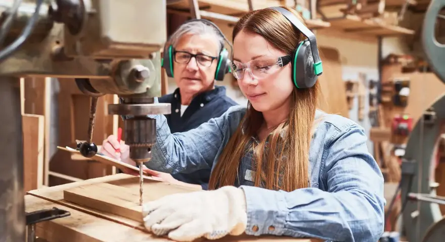 Junge Frau lernt an einer Bohrmaschine, während ein älterer Mann zusieht. Eine gute Ausbildung, welche Ausbildungen dauern 2 Jahre, ist hier vielleicht der Anfang.