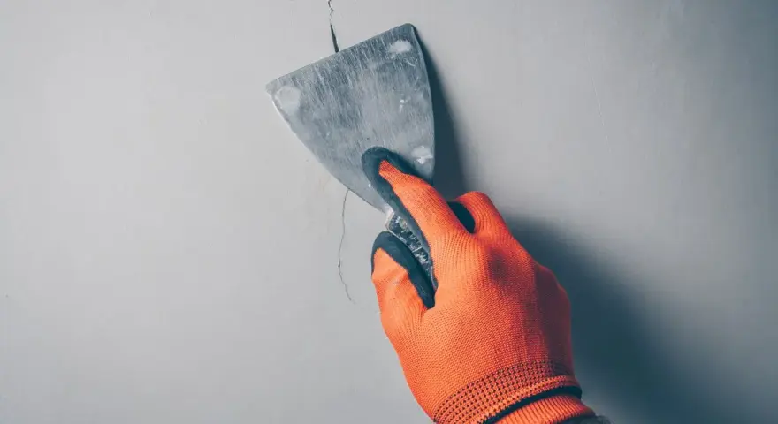 Ręka w pomarańczowej rękawicy zacierająca pęknięcie w ścianie szpachlą. Idealne rozwiązanie, czym wypełnić głębokie pęknięcia w ścianie.