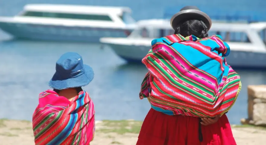 Zwei cholitas escaladoras, gekleidet in bunte Ponchos und Hüte, blicken auf Boote auf einem See.