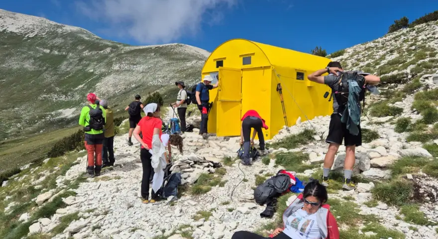 Gruppo di escursionisti vicino a un giallo blockhaus Majella, su un sentiero roccioso con montagne verdi sullo sfondo.