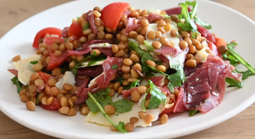 Un'appetitosa insalata di lenticchie con pomodorini, scaglie di formaggio e prosciutto crudo, guarnita con rucola fresca.