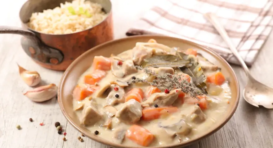 Un plat de blanquette de veau crémeuse avec des carottes, des champignons et des herbes, servi avec du riz dans une casserole en cuivre.