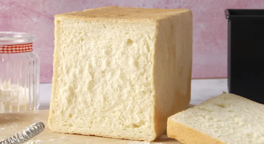 Un pain de mie moelleux, parfait pour une recette facile. Un pot de farine et un moule à pain sont à proximité.