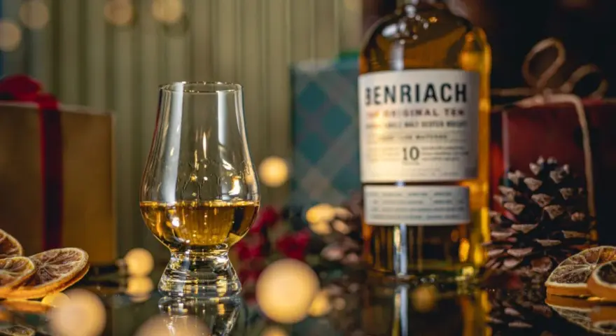 Szklanka z bursztynowym single malt whisky Benriach 10, obok butelki i świątecznych dekoracji.