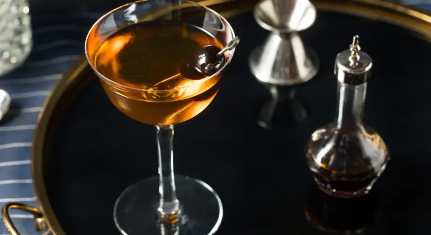 Elegancki koktajl w kolorze calvados, z wiśnią, obok kieliszka do mierzenia i buteleczki z bitters. Idealny do deserów.
