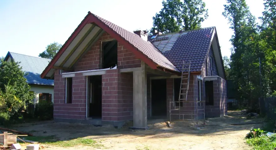 Dom w stanie surowym otwartym, z czerwonej cegły, z częściowo położonym dachem. Wokół plac budowy.