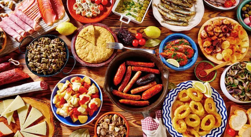 Tapas co to? Obficie zastawiony stół pełen hiszpańskich przekąsek: kiełbaski, krewetki, kalmary, owoce morza, sery, wędliny i tortilla.