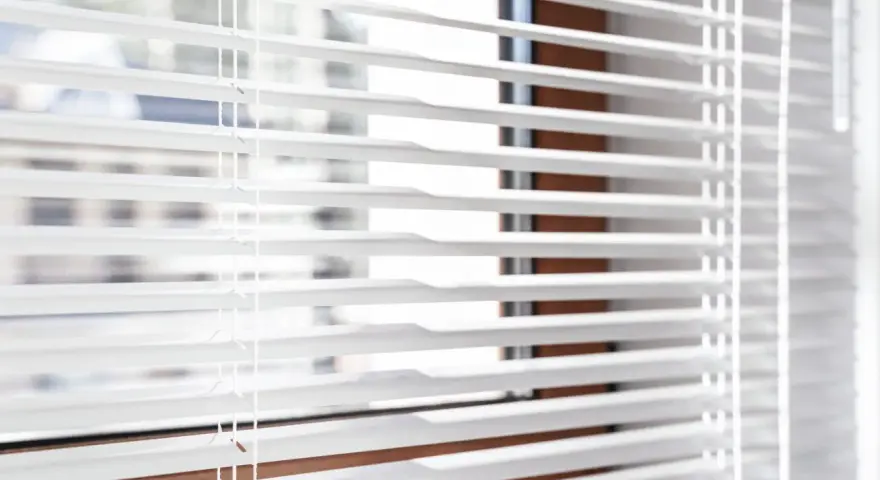Close-up of white horizontal blinds with visible cords, showing the slats and a hint of the window frame.