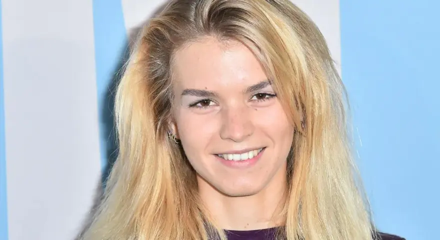Uśmiechnięta Marta Staśmierska z długimi blond włosami, w fioletowej bluzce, na tle niebiesko-białych pasów.