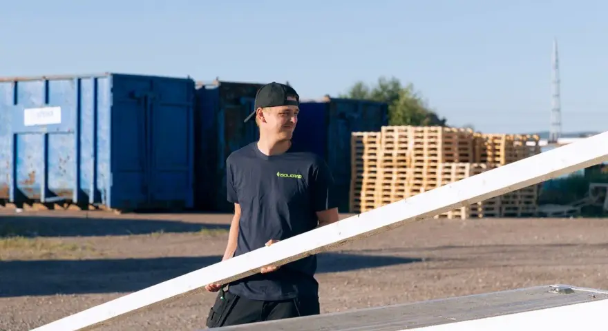 Man i mörkblå t-shirt med "ISOLERAB" logga bär en träskiva. Bakom honom syns blå containrar och träpallar.