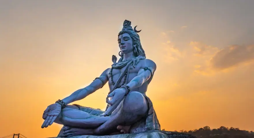 Eine riesige Statue von Shiva sitzt in meditativer Haltung vor einem warmen Sonnenuntergang.