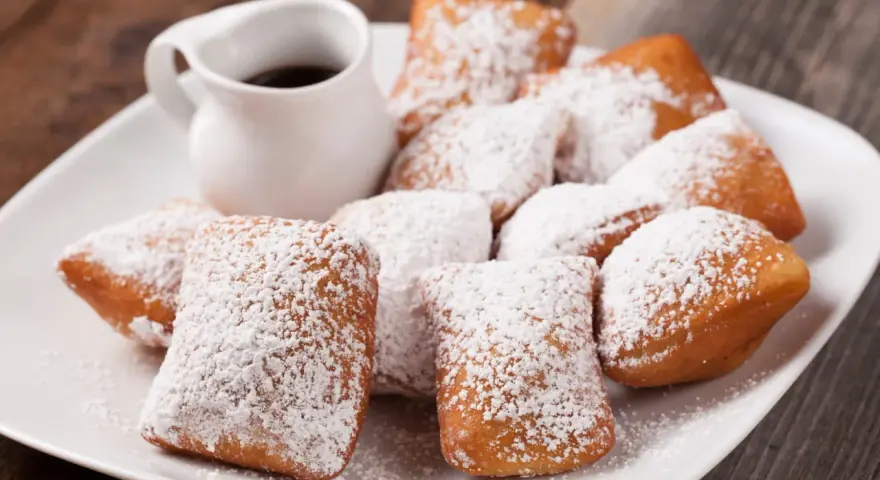Une assiette de beignets saupoudrés de sucre glace, parfaits pour une recette pâte à beignets salés. Un petit pichet de sauce est à côté.