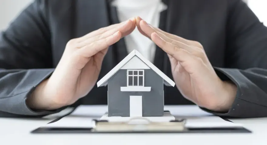 Mains protégeant une maquette de maison, symbolisant la **garantie de bon fonctionnement** et la sécurité de votre investissement immobilier.