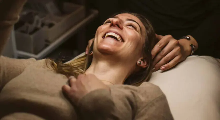 A woman laughs joyfully during a holistic facial treatment, her head tilted back as hands gently support her.