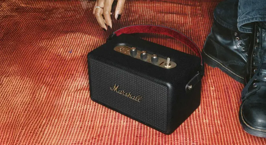 En svart Marshall Kilburn-högtalare på en röd, randig matta. En hand med ring och nagellack sträcker sig mot den.