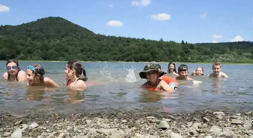 Dzieci pluskają się w jeziorze, świetny pomysł na wakacje! W tle zielone wzgórza i błękitne niebo.
