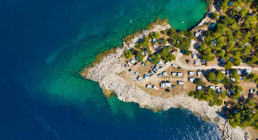 Widok z lotu ptaka na kemping Riviera Makarska. Kamieniste wybrzeże, turkusowa woda i zielone drzewa otaczają liczne przyczepy kempingowe i samochody.