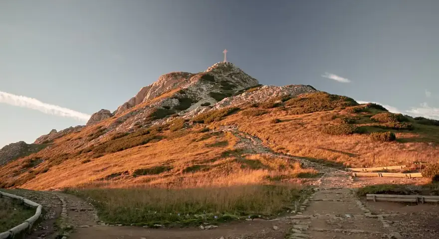 Giewont z krzyżem na szczycie, oświetlony zachodzącym słońcem. Ścieżka prowadzi w górę, a w oddali widać chmurę. Wyobraź sobie, że idziesz tam z dzieckiem.