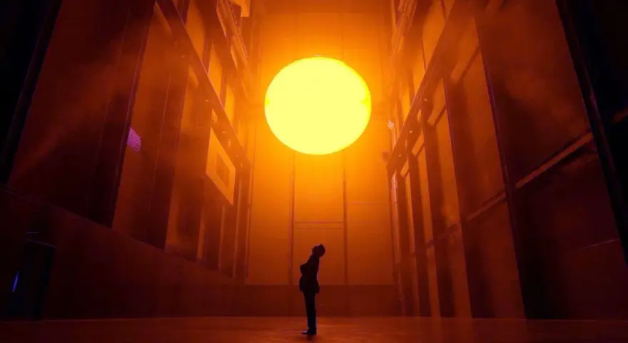 Une personne contemple un soleil artificiel dans l'installation "The Weather Project" d'Olafur Eliasson, baignant l'espace d'une lumière orangée intense.