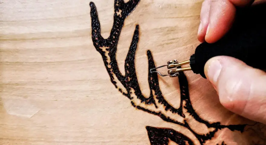 Dettaglio di un disegno a pirografia su legno: una mano guida le punte pirografo per creare le corna di un cervo.