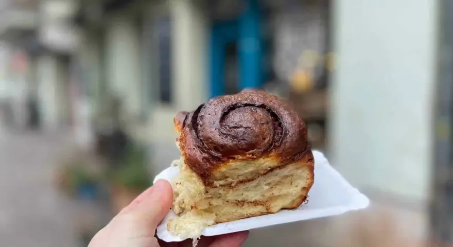 Hand hält ein saftiges Zimtschnecken-Stück vor einer unscharfen Berliner Kulisse.