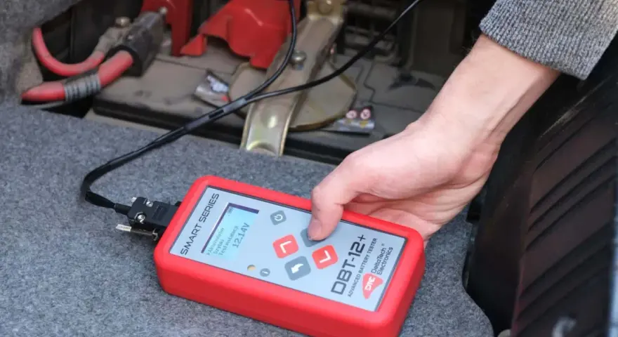 Ręka trzyma tester akumulatora DBT-12+, pokazujący 12.14V. To prosty sposób, jak sprawdzić akumulator w samochodzie.