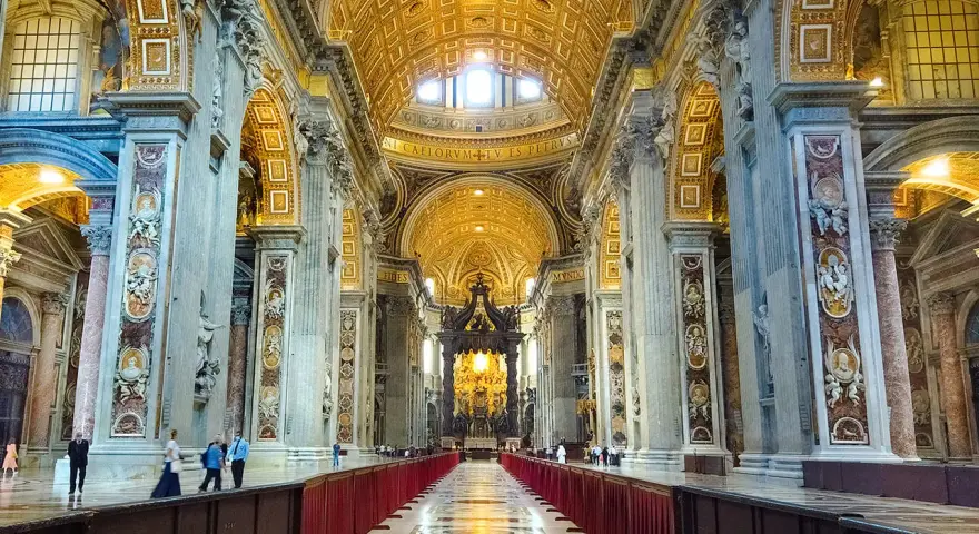 Interior of a grand basilica in Rome, with ornate golden ceilings, marble columns, and a long aisle lined with red velvet.