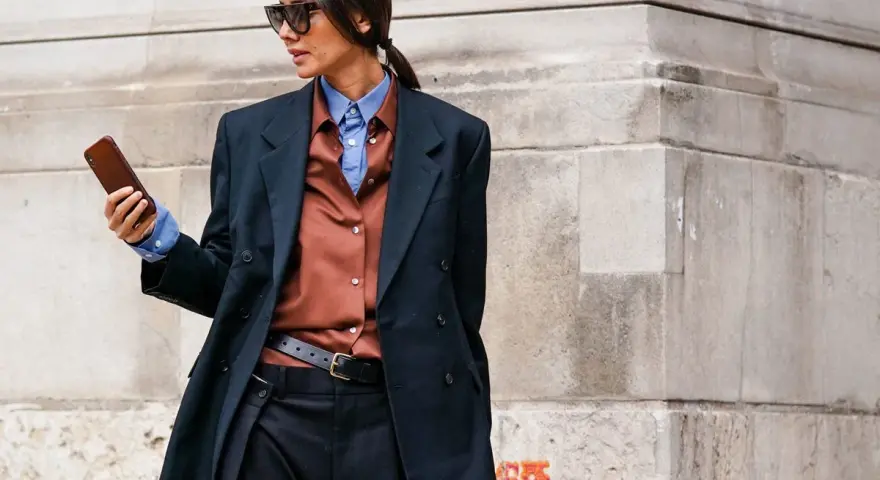 Mujer con estilo combinando traje negro con camisa marrón y azul, gafas de sol y móvil en mano.