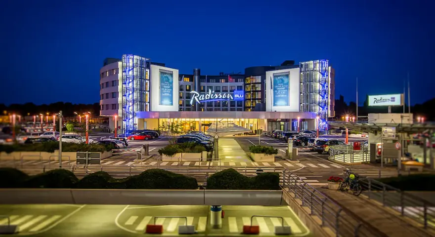 Nowoczesny hotel Radisson Blu Hamburg Airport nocą, z podświetlanym logo i parkingiem pełnym samochodów.