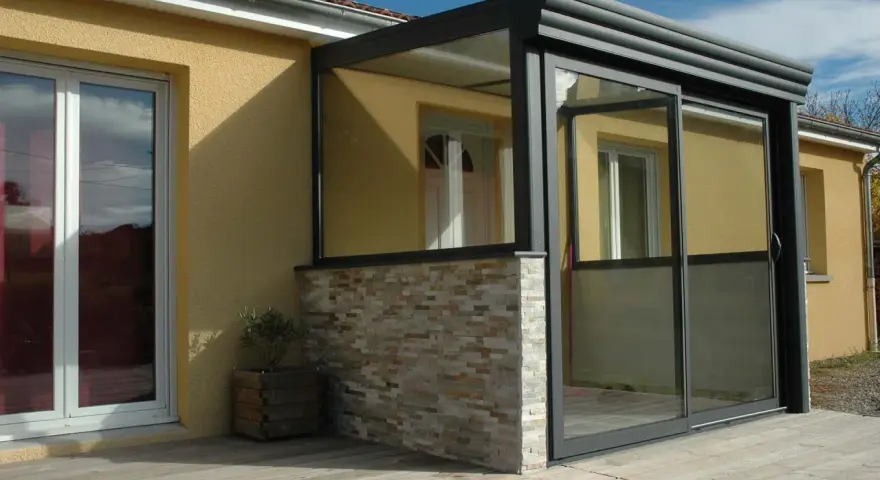 Une véranda moderne avec murs en pierre et grandes baies vitrées permet une entrée directe dans le séjour.