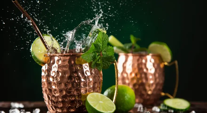 Dwa miedziane kubki z orzeźwiającym Moscow Mule, ozdobione limonką i miętą, z bryzgami wody i kostkami lodu.