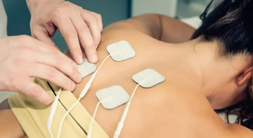 Fizjoterapeuta umieszcza elektrody na plecach pacjentki, przygotowując do zabiegu TENS. Należy pamiętać o przeciwwskazaniach do jego stosowania.