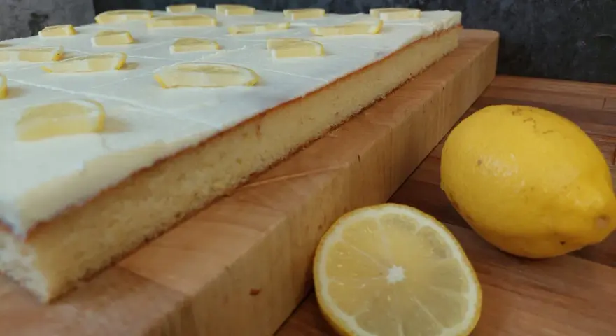 Ein saftiger sizilianischer Zitronenkuchen, bedeckt mit weißer Glasur und Zitronenscheiben, daneben frische Zitronen.