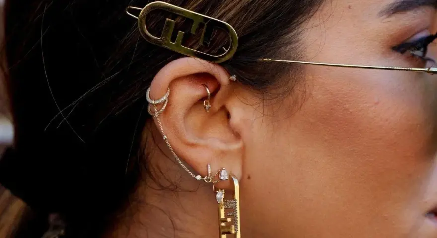 Ucho ozdobione wieloma kolczykami, w tym forward helix piercing, z błyszczącymi dodatkami i złotą spinką z logo.
