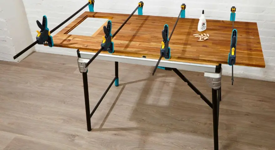 Holzplatte wird mit Zwingen fixiert, um einen Tisch selber bauen. Kleber und Dübel liegen bereit.