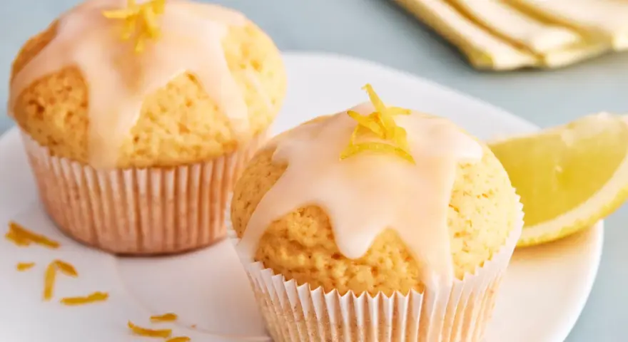 Zwei einfache Zitronenmuffins mit Zuckerguss und Zitronenschale garniert, daneben ein Zitronenschnitz.