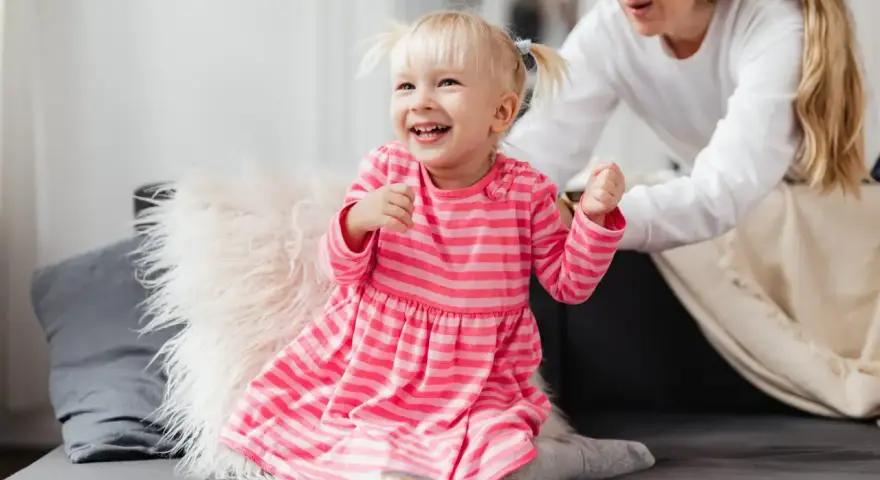 Uśmiechnięta dziewczynka w różowej sukience, która często mówi "kocham cię", bawi się z mamą na kanapie.