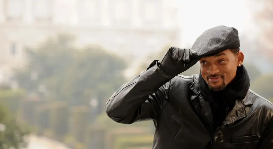 Will Smith, con un berretto uomo, giacca di pelle e sciarpa, saluta con un sorriso.