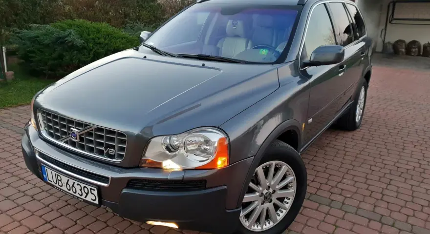 Szary Volvo XC90 z silnikiem V8, zaparkowany na podjeździe z kostki brukowej, otoczony zielenią.