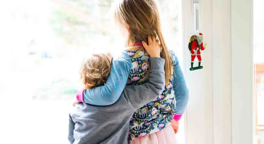 Zwei Kinder umarmen sich vor einem Fenster. Das jüngere Kind, das Angst hat, wird vom älteren Kind getröstet. Ein Weihnachtsmann-Anhänger hängt an der Tür.